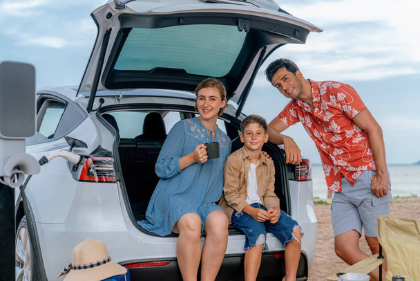 Viaggio di vacanza in famiglia viaggiando in spiaggia con auto elettrica, felice famiglia ricaricare auto EV, godendo di caffè campeggio all'aperto. Viaggi in mare e auto eco-friendly per un ambiente pulito. Perpetuo - Foto, immagini