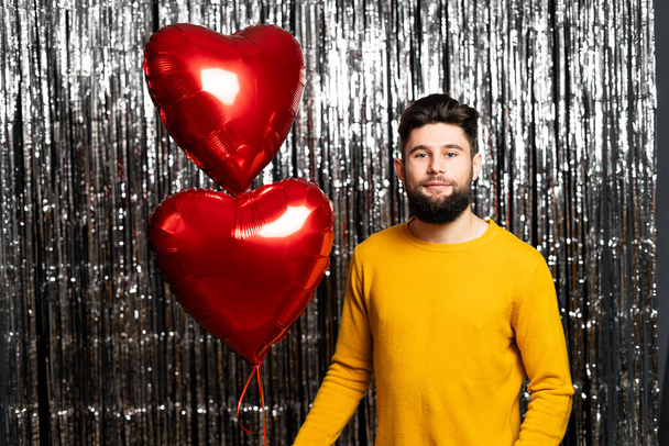 Slavíme Valentýna. Hezký stylový muž s červenými balónky ve tvaru srdce, smějící se na pozadí. Dovolenková párty. Radostný model pózování. Kopírovat prostor - Fotografie, Obrázek