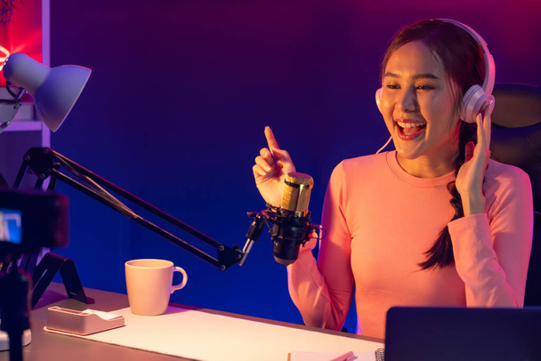 Genç ve güzel Asyalı yayıncının sunucu kanalı, dinleyicilerle sosyal medyada müzik dinliyor, pastel kulaklık takıyor, kapalı alanda neon ışıklı stüdyoda şarkı söylemek için mikrofon kullanıyor. Taktik. - Fotoğraf, Görsel
