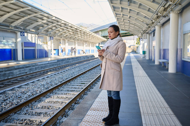 Портрет повної довжини впевненої молодої жінки в теплому зимовому одязі, перевірка мобільного додатку на її смартфоні, онлайн бронювання квитків, стоячи на відкритому повітрі на залізничній платформі вокзалу - Фото, зображення