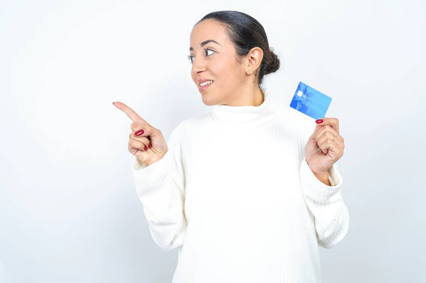 Ciekawy uśmiech piękny arabski kobieta pozowanie na białym tle pokazujące plastikowy bank pokazujący palec copyspace - Zdjęcie, obraz
