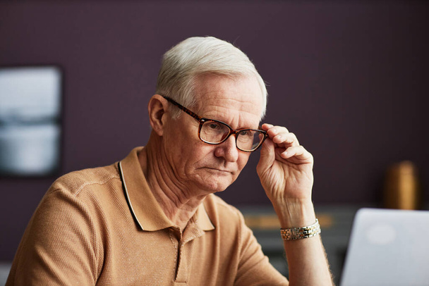 Komoly, szemüveges idősebb férfi, aki a laptopon végzett online munkájára koncentrál a szobában. - Fotó, kép