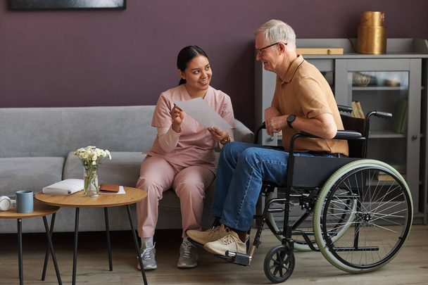 Junge Pflegerin erklärt Senioren mit Behinderung die Art der Bezahlung von Rechnungen, während sie im Zimmer sitzen - Foto, Bild