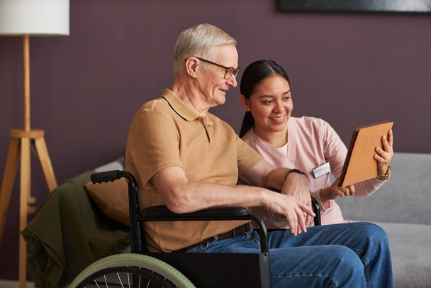 Молодий волонтер навчає старшого чоловіка з інвалідністю користуватися цифровим планшетом, сидячи вдома - Фото, зображення