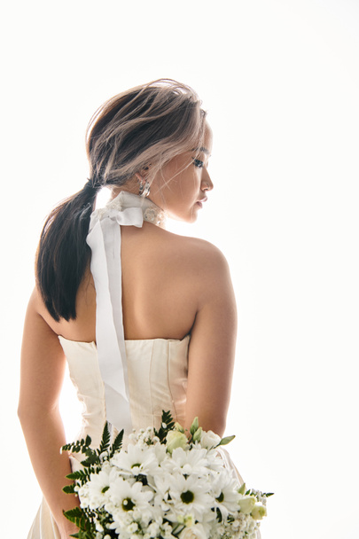 verleidelijke jonge bruid in witte jurk met boeket bloemen achter haar rug op lichte achtergrond - Foto, afbeelding