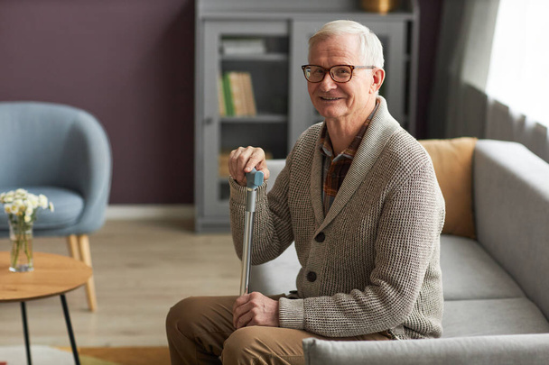 Portrait d'un homme âgé souriant à la caméra tout en se reposant sur un canapé dans le salon - Photo, image