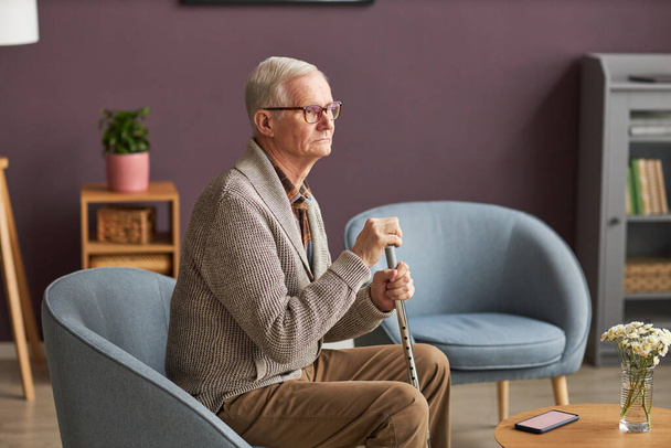 Uomo anziano con bastone seduto sulla poltrona con vista pensierosa, trascorre del tempo a casa - Foto, immagini