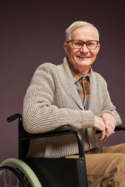 Retrato de un joven anciano con discapacidad sentado en silla de ruedas y sonriendo a la cámara sobre fondo gris - Foto, Imagen