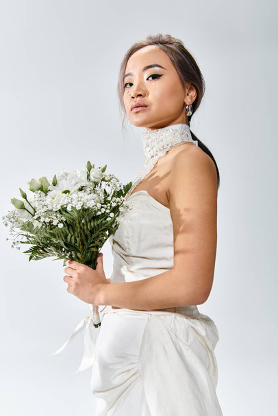 привлекательная азиатская невеста в элегантном наряде с белыми цветами букет глядя боком на камеру - Фото, изображение