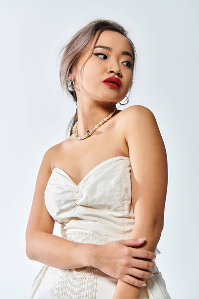 alluring asian woman in her 20s with red lips and pearl necklace look to side on white background - Foto, Bild