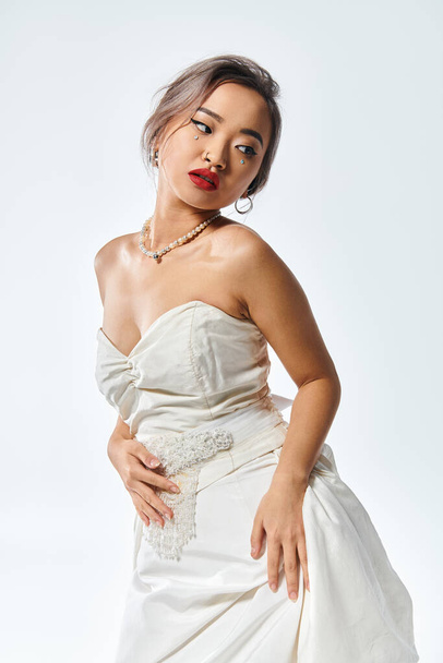 attractive asian bride with red lipstick and pearl necklace looking behind back on white background - Zdjęcie, obraz