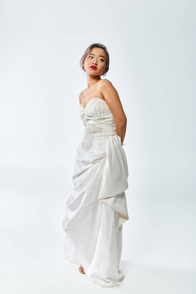 mooie aziatische bruid met rode lippen en parel ketting poseren en op zoek naar kant op witte achtergrond - Foto, afbeelding