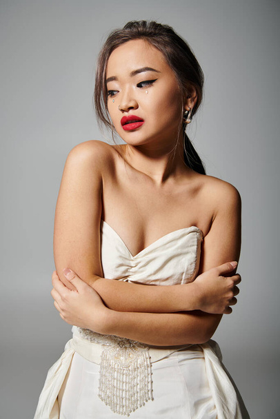 alluring asian young girl with pearl necklace and red lipstick hug herself on grey background - Photo, Image