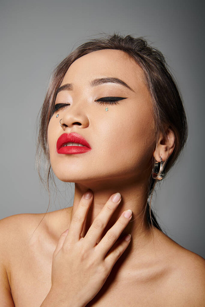 graceful asian girl in her 20s with closed eyes and red lipstick hug to neck on grey background - Photo, Image