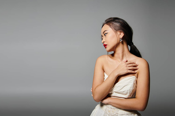 seductora mujer con labios rojos cubriendo el escote y mirando hacia un lado contra fondo gris - Foto, imagen