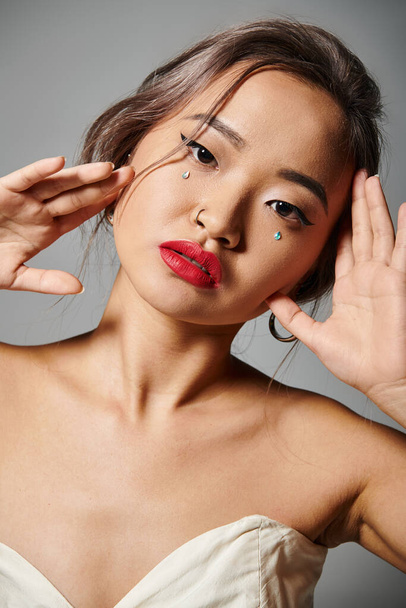 ritratto di bella donna asiatica con labbra rosse che si tengono le mani intorno al viso su sfondo grigio - Foto, immagini
