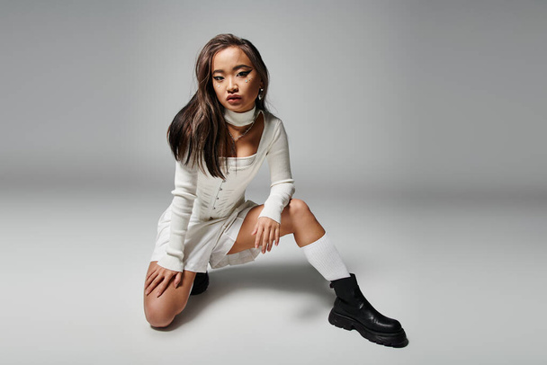 daring asian young girl in stylish outfit crouched down against grey background - Photo, Image
