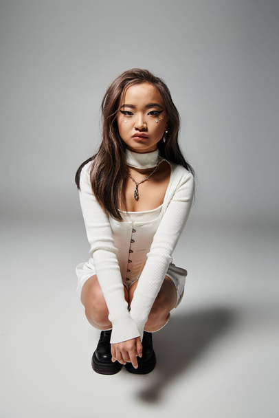 pretty asian young girl in white stylish outfit crouched down against grey background - Zdjęcie, obraz