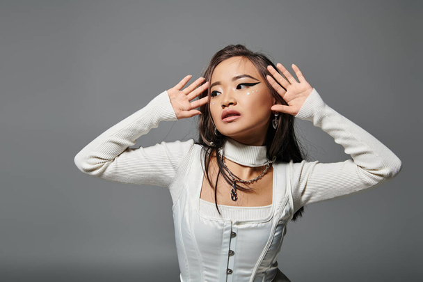 заманчивая азиатская женщина в возрасте 20 лет смотрит в сторону и позирует руками на сером фоне - Фото, изображение