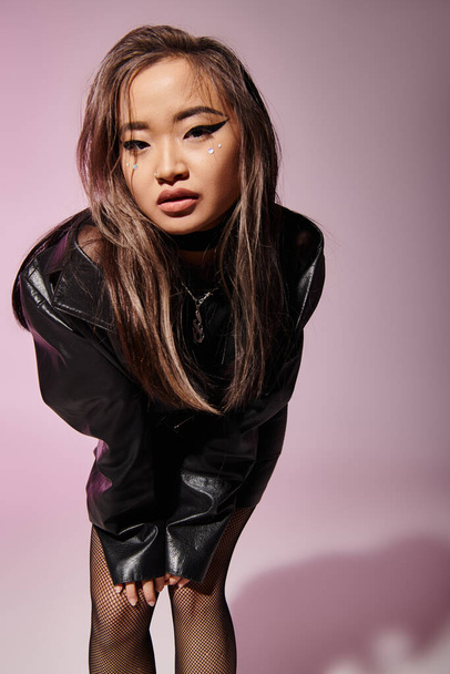 chica joven asiática en traje de cuero negro con maquillaje pesado inclinado hacia adelante sobre fondo lila - Foto, imagen
