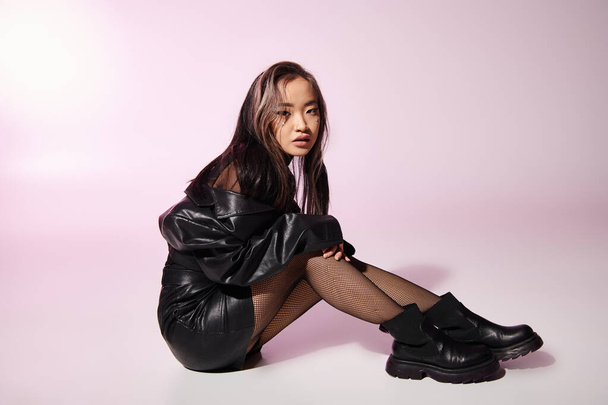 lovely asian woman in black leather outfit with heavy makeup sitting sideways on lilac background - Photo, Image