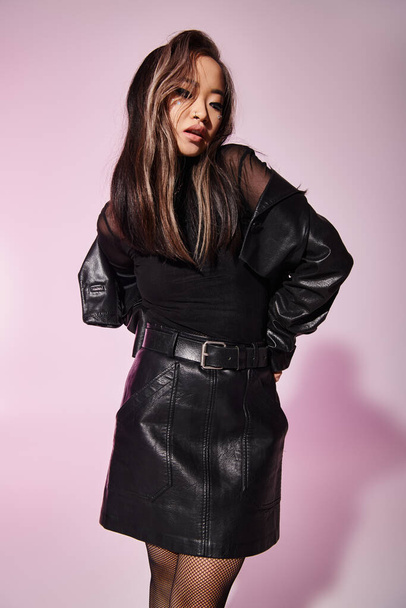 очаровательная азиатская молодая женщина в черной кожаной одежде с тяжелым макияжем стоя и глядя вниз - Фото, изображение