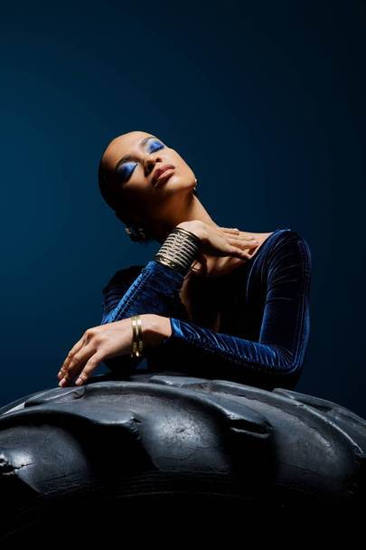 Young African American woman wearing a blue dress sits gracefully on top of a tire in a studio setting. - Photo, Image