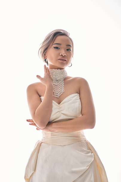 encantadora mujer joven asiática con cabello castaño en vestido elegante blanco posando con las manos - Foto, Imagen