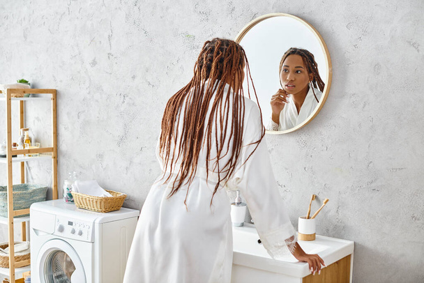 Afrikkalainen amerikkalainen nainen kylpytakissa ja afro-punoksissa harjaa hampaansa peilin edessä modernissa kylpyhuoneessa.. - Valokuva, kuva