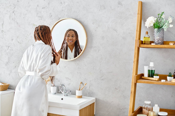 Μια Αφρο-Αμερικανίδα με αφρο-πλεξούδες στέκεται στο μοντέρνο μπάνιο της θαυμάζοντας την αντανάκλασή της στον καθρέφτη.. - Φωτογραφία, εικόνα