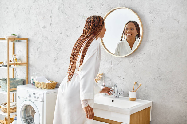 Mulher afro-americana em roupão de banho com tranças afro fica na frente da máquina de lavar roupa moderna no elegante banheiro. - Foto, Imagem