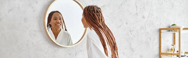 Uma mulher afro-americana com tranças afro olha para seu reflexo em um espelho em um banheiro moderno, contemplando beleza e higiene. - Foto, Imagem