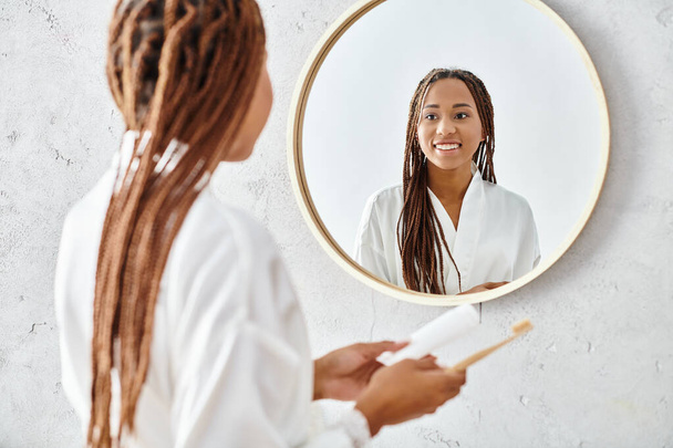 Una mujer afroamericana con trenzas afro se para frente a un espejo en un baño moderno, con una bata de baño. - Foto, imagen