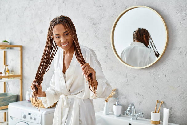 African American woman with afro braids in bath robe, stands in front of modern bathroom sink, admiring her reflection. - Fotoğraf, Görsel