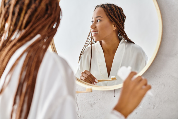 An African American woman in a bathrobe with afro braids brushes her teeth in front of a mirror in a modern bathroom. - Zdjęcie, obraz