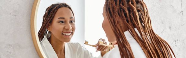 Une Afro-Américaine avec des tresses afro se brosse les dents dans une salle de bain moderne, se concentrant sur sa beauté et son hygiène. - Photo, image
