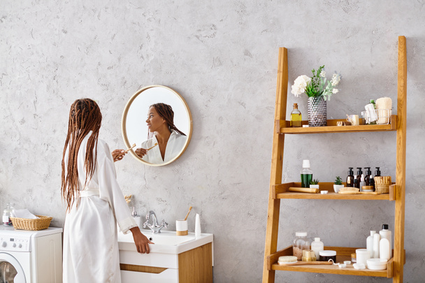 Stylowa Afroamerykanka w szlafroku z afro warkoczami stojąca przed nowoczesnym umywalką w łazience. - Zdjęcie, obraz