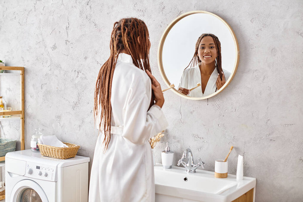 Een Afro-Amerikaanse vrouw met afro vlechten staat voor een spiegel in een moderne badkamer terwijl ze een badjas draagt. - Foto, afbeelding