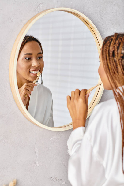 Uma mulher afro-americana com tranças afro escova os dentes em um banheiro moderno, vestindo um roupão de banho. - Foto, Imagem