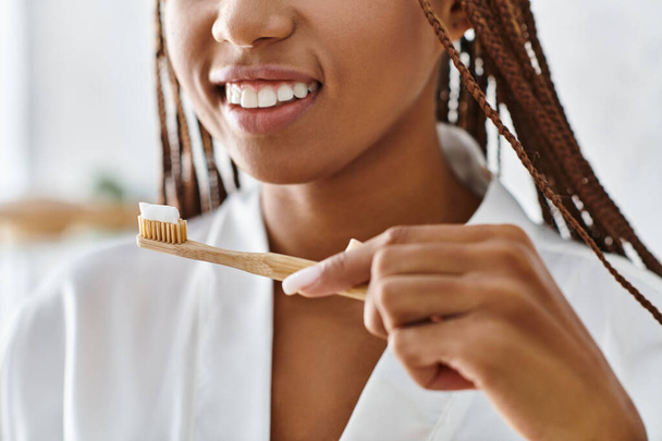 Μια Αφρο-Αμερικανίδα με μπουρνούζι με αφρο-πλεξούδες βουρτσίζει τα δόντια της σε ένα μοντέρνο μπάνιο για ομορφιά και υγιεινή.. - Φωτογραφία, εικόνα