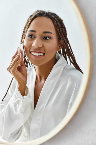 Une Afro-Américaine avec des tresses afro utilisant du coton avec du toner devant un miroir dans une salle de bain moderne tout en portant un peignoir de bain. - Photo, image