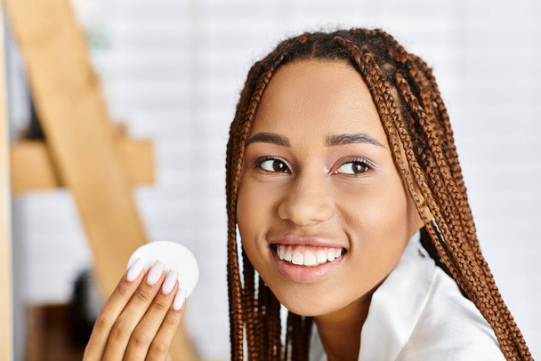Africano mulher americana com tranças afro em roupão de banho, segurando um objeto branco em sua mão em um banheiro moderno. - Foto, Imagem