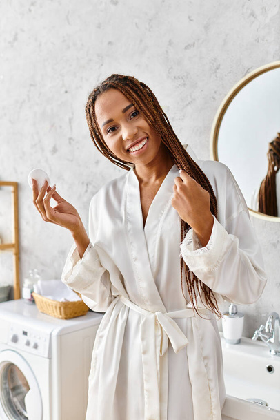 Bornozlu Afro-Amerikan bir kadın modern bir banyoda pamuk tutarak güzellik ve hijyene odaklanıyor.. - Fotoğraf, Görsel