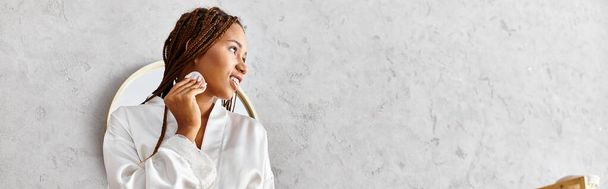 Αφρο-Αμερικανίδα με αφρο-πλεξούδες σε μπουρνούζι κρατώντας βαμβακερό μαξιλάρι μπροστά από έναν καθρέφτη σε ένα μοντέρνο μπάνιο. - Φωτογραφία, εικόνα