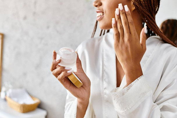 Mujer afroamericana con trenzas afro en albornoz sosteniendo un frasco de crema frente a su cara en un baño moderno. - Foto, imagen