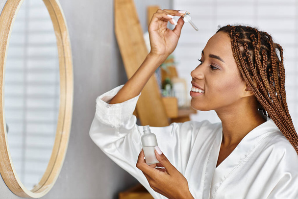 Afroamerikanische Frau mit Afro-Zöpfen, die eine Flasche mit Serum vor dem modernen Badezimmerspiegel hält. Schönheit und Hygiene-Routine. - Foto, Bild