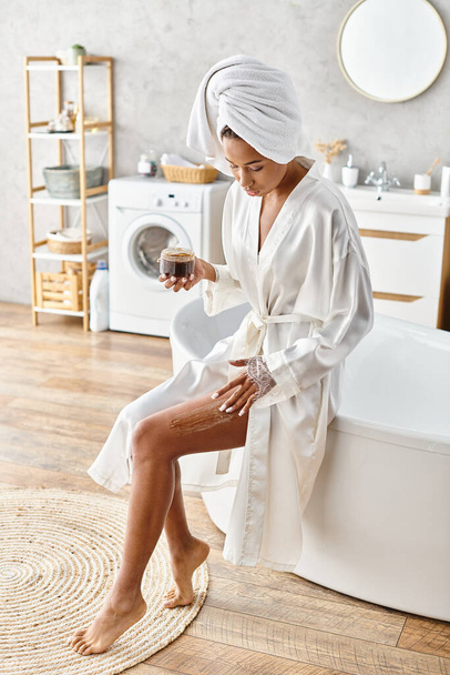 Eine afroamerikanische Frau im Bademantel, Peeling-Körper mit Peeling, während sie in einer Badewanne in ihrem modernen Badezimmer sitzt. - Foto, Bild