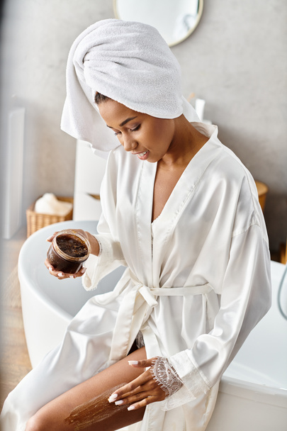 Una mujer afroamericana en un albornoz se relaja con un exfoliante de café en su moderno baño, abrazando un momento de calma y belleza. - Foto, imagen