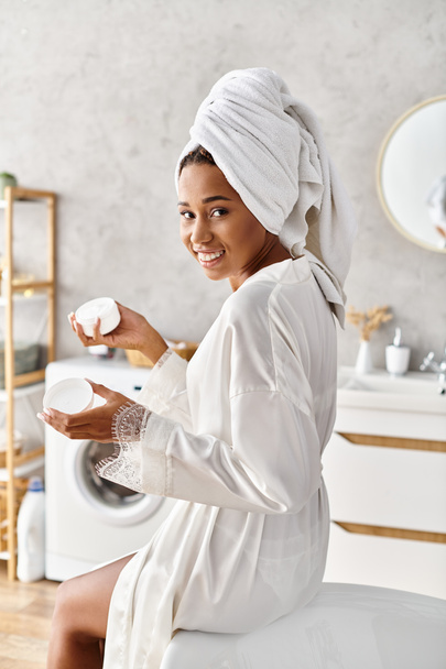 An African American woman in a bathrobe with a towel wrapped around her head, holding jar with cream - Photo, Image