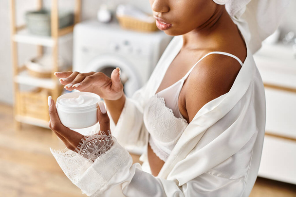 Femme afro-américaine avec des tresses afro en robe blanche tenant pot de crème dans la salle de bain moderne. - Photo, image
